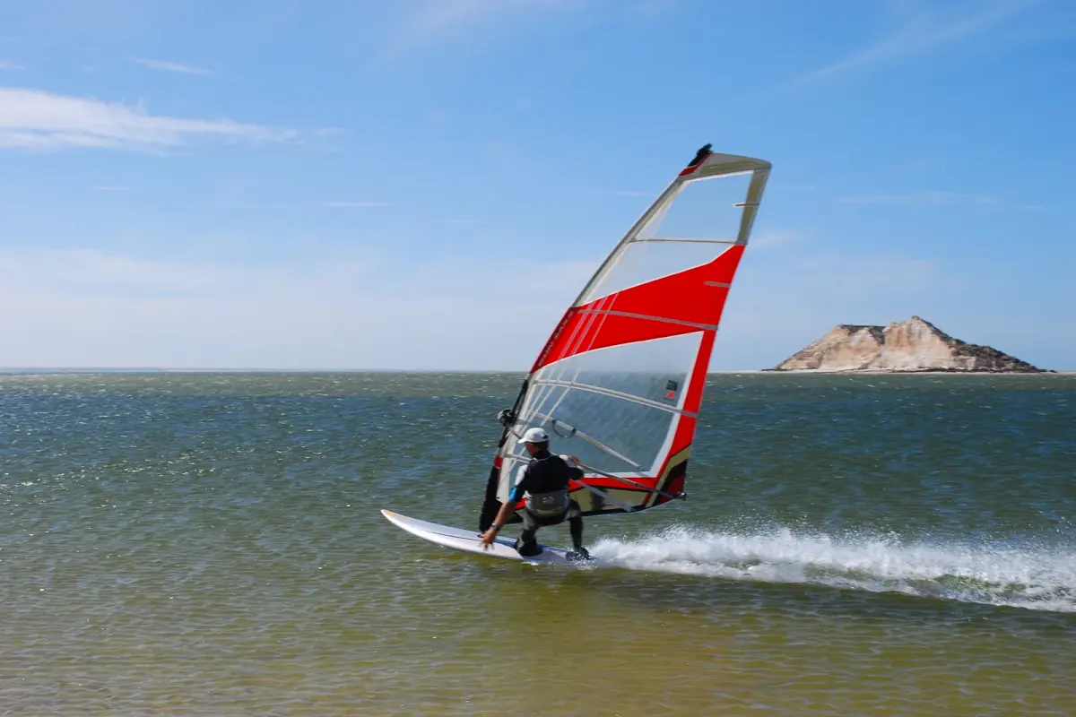 Un windsurfeur en combinaison file sur une eau verte et calme, voile rouge et blanche bordée, devant l'île du Dragon, un rocher clair posé sur la lagune de Dakhla