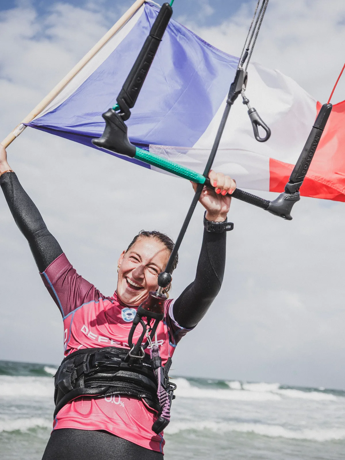 Capucine Delannoy en lycra de comp&eacute;tition, bras lev&eacute;s au bord de l'eau, tenant un drapeau fran&ccedil;ais accroch&eacute; &agrave; sa barre de kite