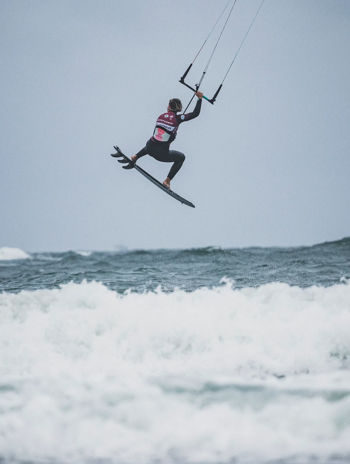 Capucine Delannoy en plein saut au-dessus de l'&eacute;cume, pieds nus pos&eacute;s sur sa planche de surf sans straps, une main sur la barre