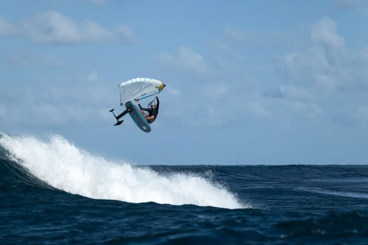Rider en wing foil retourn&eacute; tr&egrave;s haut au-dessus d'une vague, aile Duotone blanche et cyan &agrave; bord d'attaque jaune tenue &agrave; bout de bras, planche sombre et foil apparents