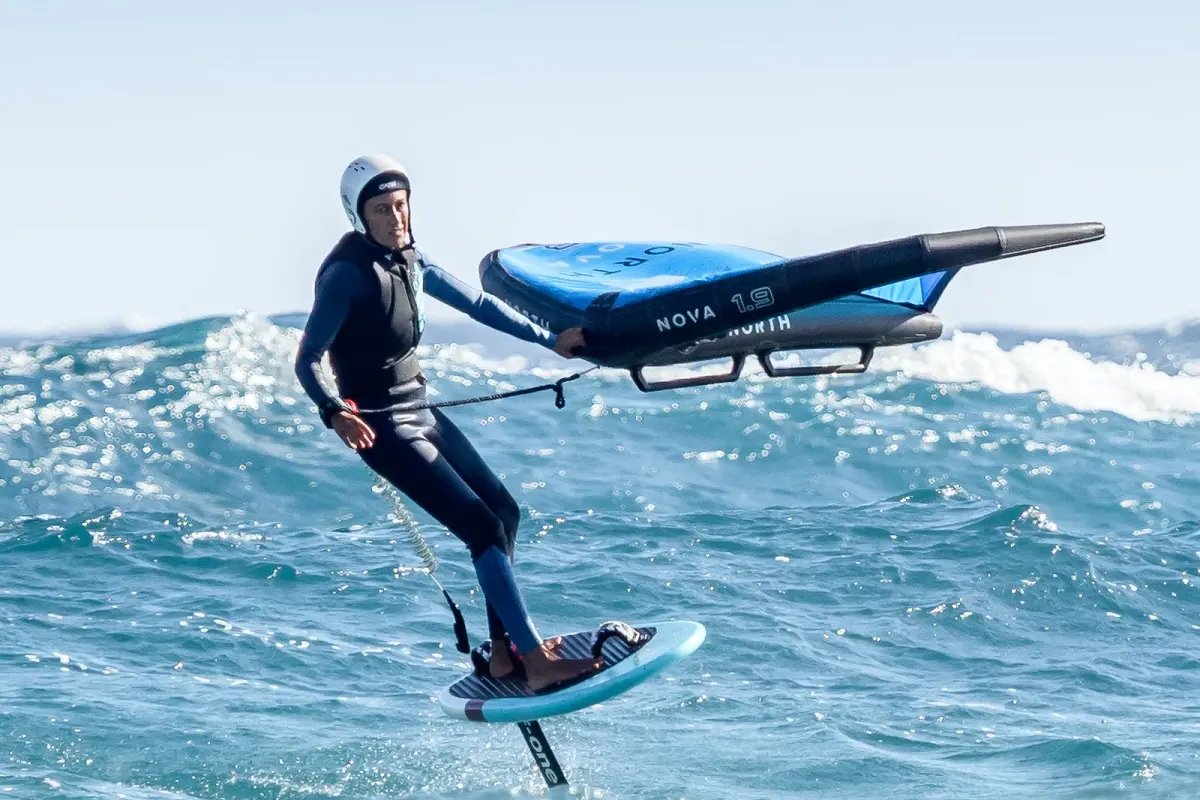 Un wingfoiler casqué vole sur son foil au-dessus d'une mer agitée, l'aile bleue tenue d'une main, une vague qui déferle derrière lui
