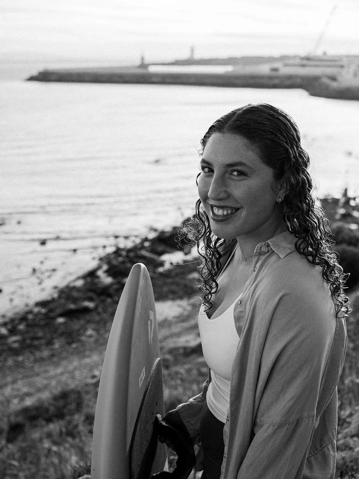 Portrait en noir et blanc de Nia Suardiaz, souriante, une planche de wingfoil sous le bras, au bord de la mer à Tarifa