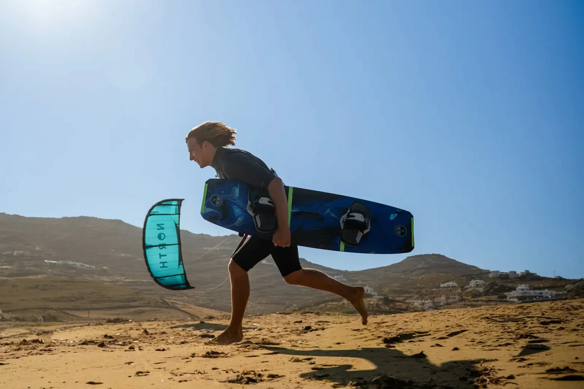 Un kitesurfeur court sur une dune de sable avec une planche North Atmos sous le bras, son aile pos&eacute;e derri&egrave;re lui