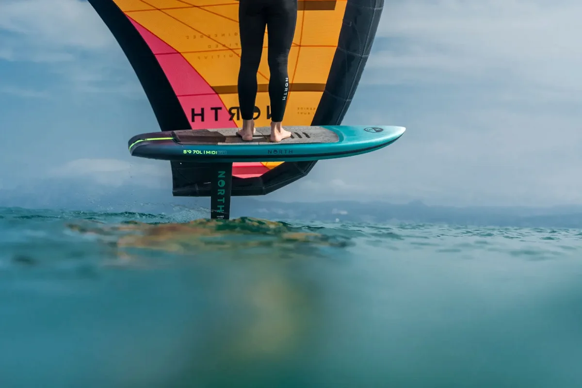 Un rider en wingfoil vole au-dessus de l'eau sur une planche North Midi Pro turquoise, sous une aile orange et rose