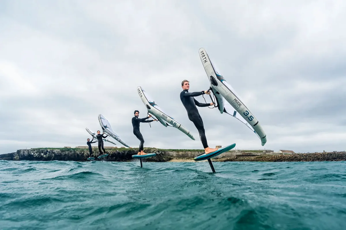 Trois riders en wingfoil sous des ailes North blanches, devant une digue de pierre et une mer verte