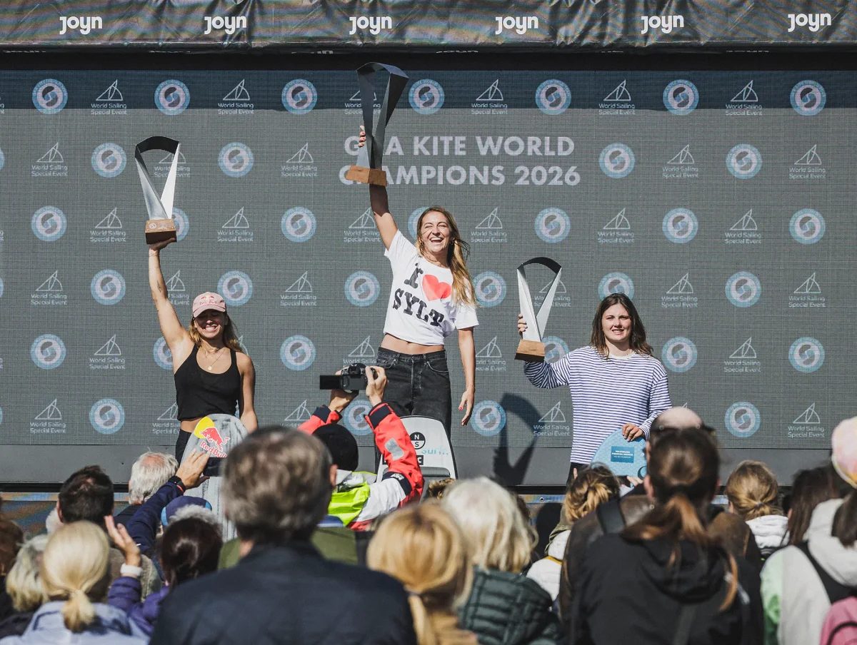 Trois rideuses sur le podium, troph&eacute;es lev&eacute;s, devant un &eacute;cran affichant &laquo; GKA Kite World Champions 2026 &raquo;, public au premier plan