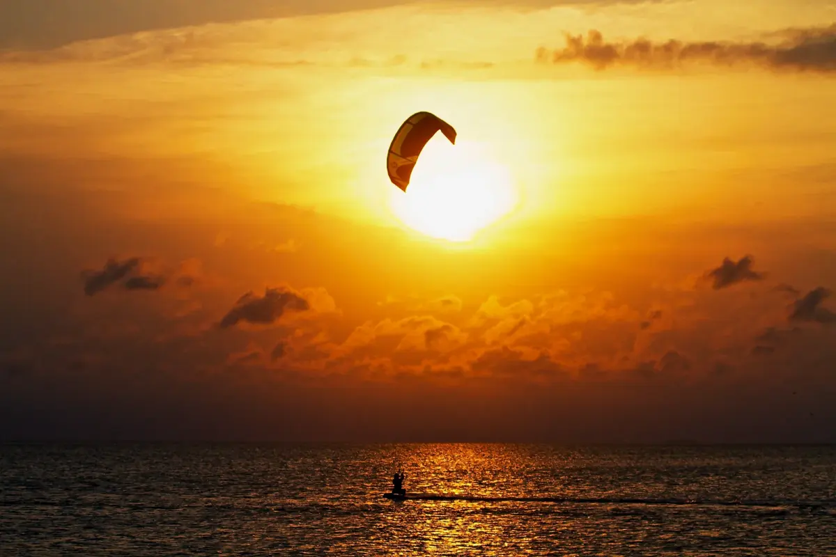 Une aile de kite en silhouette devant un soleil bas et orange, au-dessus d'un kitesurfeur qui file dans le reflet du soleil sur la mer