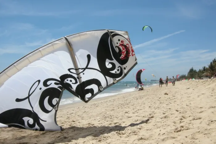 Aile de kitesurf posée à plat sur le sable, bord d'attaque et lattes gonflés au premier plan, avec d'autres kites et des riders sur la plage à l'arrière-plan