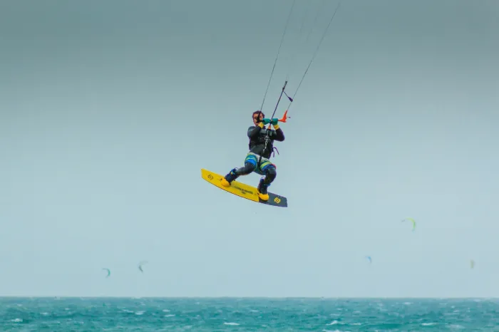 Kitesurfeur en vol à plusieurs mètres au-dessus d'une mer grise, planche aux pieds, lignes tendues vers son aile hors du cadre