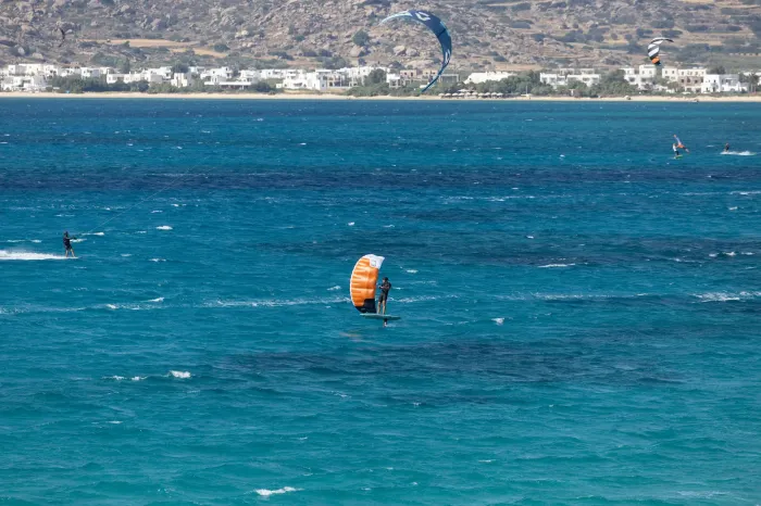 Un rider en wing foil vole au large d'une plage, un kitesurfeur le croise sur la gauche, mer bleue et vent régulier