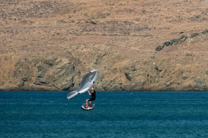 Un rider en vol sur sa planche à foil, aile grise tenue à bout de bras au-dessus de lui, devant une côte rocheuse et sèche