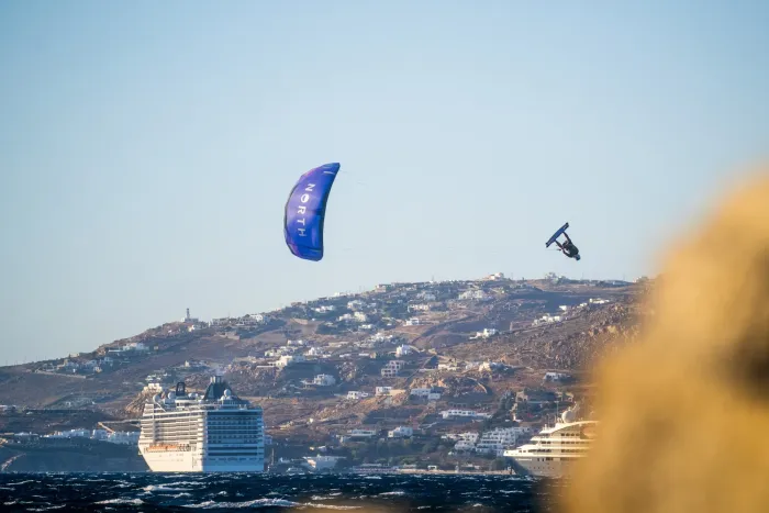 Kitesurfeur retourné en plein saut sous une aile North bleue, au-dessus d'une mer sombre et devant une côte grecque