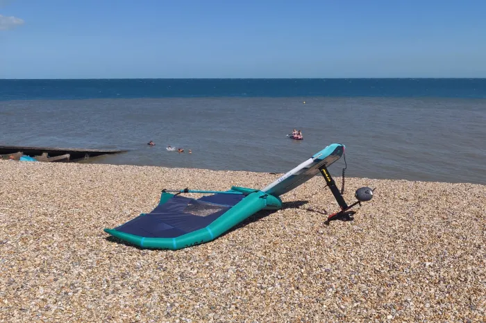 Matériel de wing foil posé sur une plage de galets, aile gonflable turquoise et bleu marine à plat, planche dressée sur son mât de foil