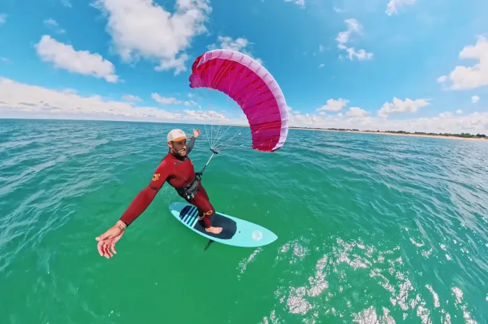 Rider en combinaison rouge sur une planche de foil, tractant une parawing rose sans boudin ni latte, au large d'une côte de sable