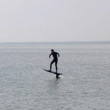 Un rider en combinaison intégrale vole sur un foil électrique au ras d'une eau plate et grise, sans aile ni pagaie pour le tracter